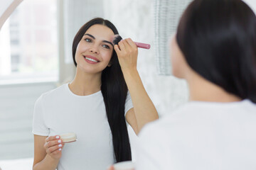 A young beautiful brunette woman at home looks in the mirror in the morning, smiles, applies makeup with a brush