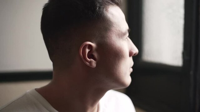 Close up of a latino young man looking out the window and giving a serious look at the camera in a white shirt