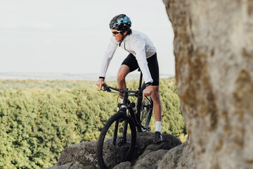 Fototapeta premium Equipped Professional Male Cyclist Riding Mountain Bike on Trail Among Rocky Terrain, Adult Man Enjoying His Extreme Hobby Standing with Bicycle on the Rock with Panoramic View