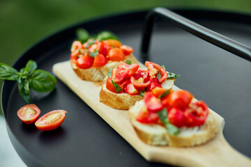 Italian bruschetta with fresh red tomatoes and basil herbs on a wooden board outdoor on the yard, backyard at home, sunlight. Classic Italy food, antipasto