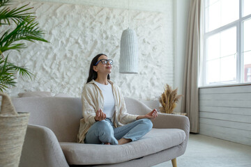 Fototapeta premium A beautiful brunette woman sits on the couch at home, rests, meditates in the lotus position, does yoga