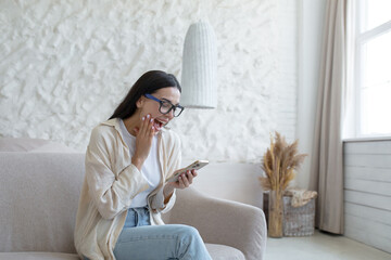 A young beautiful woman at home is sitting on the sofa in the living room, smiling joyfully,...