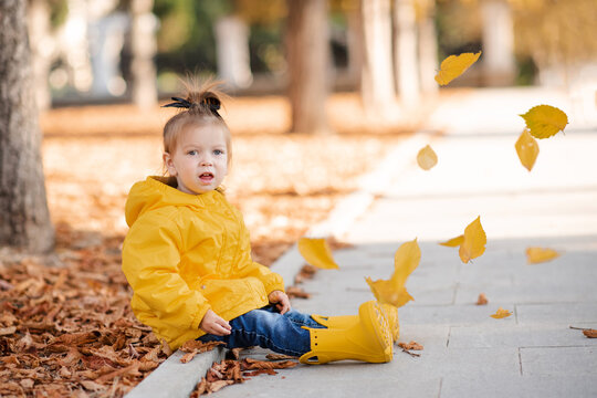 Cute funny kid girl 2-3 year old wear yellow rain jacket and rubber boots in autumn park outdoor over fallen leaves. Fall season. Childhood.