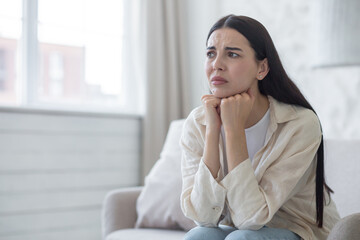Depressed woman near window sitting on sofa at home, disappointed and sad brunette grieving divorce.