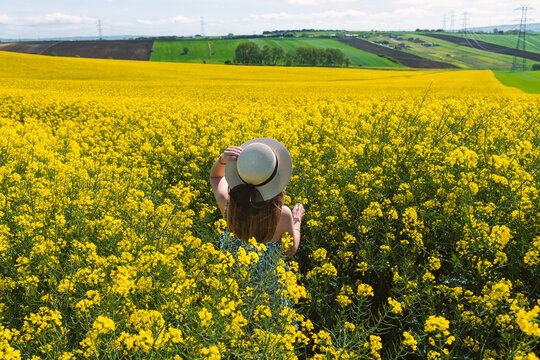 Beautiful girl with dress and hat in the Canola Fields. Blooming canola fields under a blue sky with clouds. Beautiful yellow flowers. Happy girl in nature.