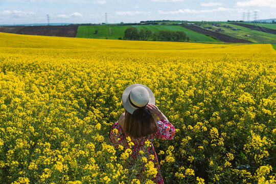 Beautiful girl with dress and hat in the Canola Fields. Blooming canola fields under a blue sky with clouds. Beautiful yellow flowers. Happy girl in nature.