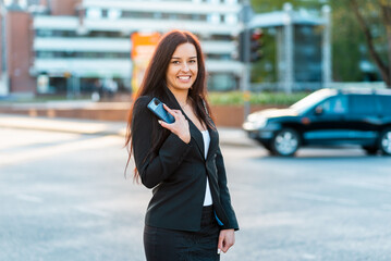 Adorable business woman with smartphone standing against street blurred building background. Fashion business photo of beautiful girl in black suite with phone.Closeup.