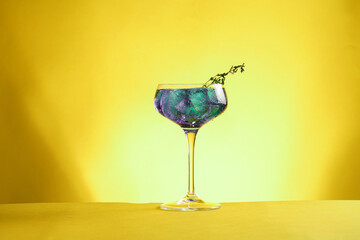 Dark blue drink in a vintage glass for sparkling wine - pea flower tea or blue curacao sirup cocktail with thyme branches on yellow backgroud. Copy space for text