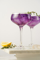 a purple drink in a vintage glass for sparkling wine - pea flower tea or blue curacao sirup cocktail with thyme branches on white background. Copy space for text