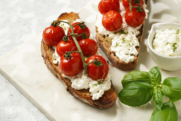 A sandwich with roasted cherry tomatoes with branch, fresh cottage cheese, green basil on a slice of whole wheat bread on a marble board on grey background
