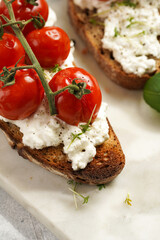 A sandwich with roasted cherry tomatoes with branch, fresh cottage cheese, green basil on a slice of whole wheat bread on a marble board on grey background