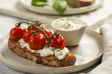 A sandwich with roasted cherry tomatoes with branch, fresh cottage cheese, green basil on a slice of whole wheat bread on a round plate on grey background
