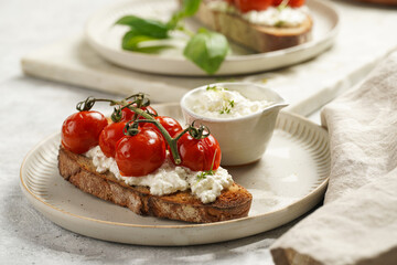 A sandwich with roasted cherry tomatoes with branch, fresh cottage cheese, green basil on a slice of whole wheat bread on a round plate on grey background