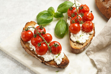 A sandwich with roasted cherry tomatoes with branch, fresh cottage cheese, green basil on a slice of whole wheat bread on a marble tray on grey background
