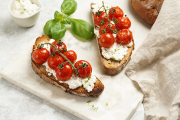 A sandwich with roasted cherry tomatoes with branch, fresh cottage cheese, green basil on a slice of whole wheat bread on a marble tray on grey background