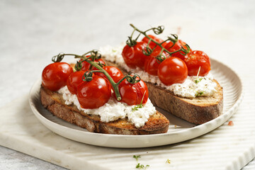 Fototapeta na wymiar a sandwich with roasted cherry tomatoes with branch, fresh cottage cheese, green basil on a slice of whole wheat bread on a round plate on marble tray on grey background