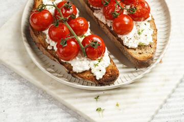 a sandwich with roasted cherry tomatoes with branch, fresh cottage cheese, green basil on a slice of whole wheat bread on a round plate on marble tray on grey background