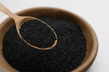 Wooden spoon of black sesame seeds above bowl on white table, closeup