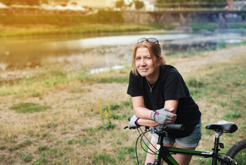 Fototapeta na wymiar A mature woman paused while riding a bicycle, takes a breath and smiles, and is about to quickly ride to the next stop. A woman rides a mountain bike on the river bank