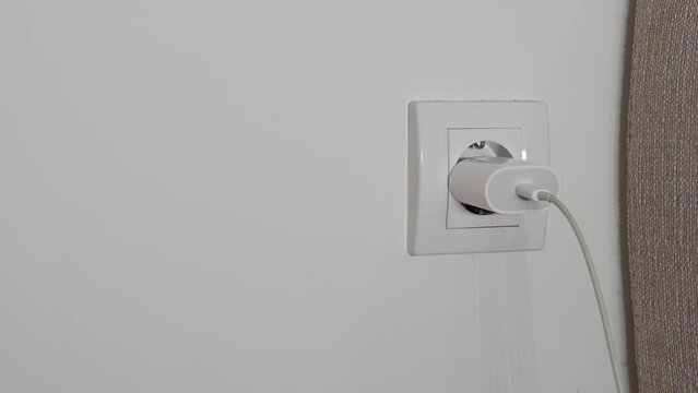 A close-up of a man's hand inserts a white power adapter to charge the phone into an outlet against a white wall.