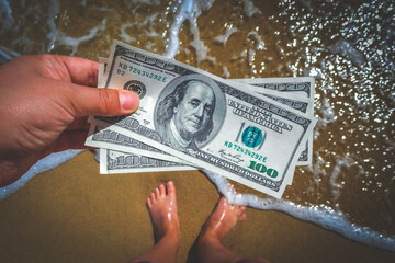 Girl holding money bill of 300 dollars on background of sea ocean waves with white foam and sand wet beach with legs foot in sand close-up. Concept finance money holiday traveling dollars vacation
