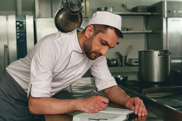Naklejka na ściany i meble Smiling chef in uniform making notes in notebook standing on kitchen