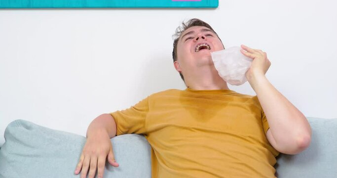Mature plump man relaxes on comfortable grey sofa giving approval sign thumb-up gesture. Guy sweats and rubs face and neck with ice pack on hot sultry day at home