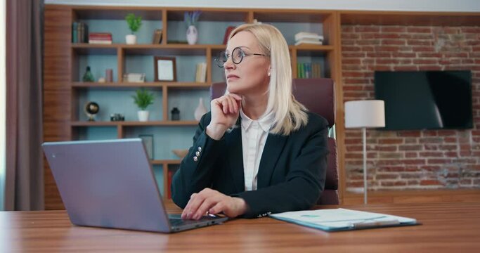 Attractive confident skilled adult blond businesswoman working on laptop and distracted for a moment from work in modern office room