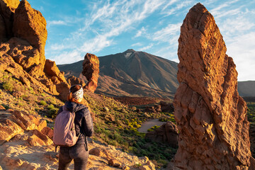 Woman with backpack hiking with scenic golden hour sunrise morning view on unique rock formation Roque Cinchado, Roques de Garcia, Tenerife, Canary Island, Spain, Europe. Pico del Teide volcano summit - obrazy, fototapety, plakaty