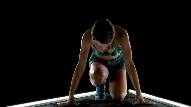 Female runner in race  start position. Girl in sportswear posing on lit track against dark background.