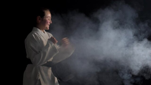 Young karate girl wearing kimono smiling and posing against black background with smoke