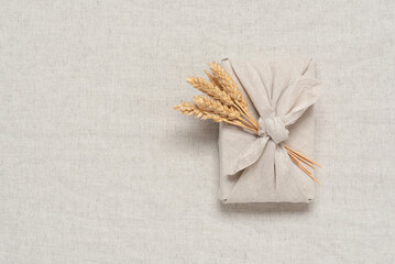Fototapeta na wymiar A gift wrapped in fabric with ears of ripe wheat. Thanksgiving gift in furoshiki style. Beige linen textile background. Top view, flat lay, selective focus.