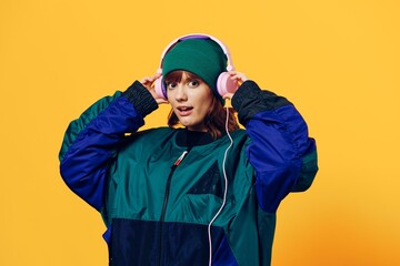 a happy, joyful woman in a green jacket and hat enjoys music standing in pink headphones on a yellow background holding them with her hands and looking at the camera