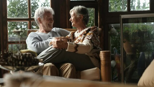 Cheerful couple of senior people enjoy leisure indoor activity together sitting on a sofa inside a winter holiday wooden home chalet. Aged man and woman smile and laugh a lot having fun. Retired life