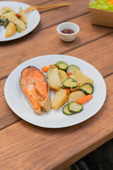 Salmon grilled fish with grilled vegetables in eco disposable tableware. Sauce container with barbecue sauce. Grilled potato, carrot, vegetable marrow. Picnic in park. Vertical photo.