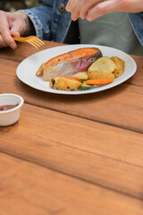 Salmon grilled fish with grilled vegetables in eco disposable tableware. Grilled potato, carrot, vegetable marrow. Man water salmon with lemon. Man eating fish. Picnic in park. Vertical photo.