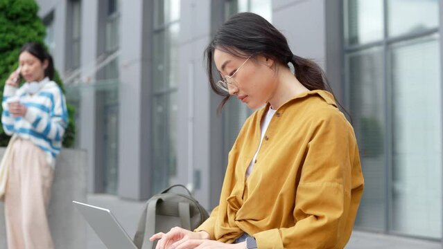 Side view of pretty chinese woman typing on wireless laptop while sitting on outdoors bench. Charming brunette standing near, drinking coffee and talking on mobile. Big city lifestyles.