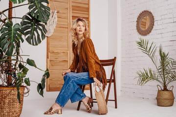 Fashionable woman wearing stylish boho outfit with suede jacket, wide leg jeans, sandals, holding trendy round wicker straw bag, posing in cozy room with green plants. Copy, empty space for text