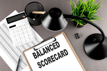 Clipboard ,chart and calculator, lamp and workspace accessories on a grey table. Top view , text BALANCED SCORECARD