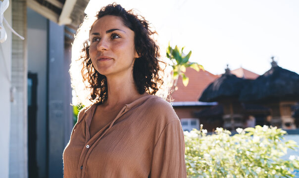 Contemplative female with curly hair thoughtful looking away thinking about weekend pastime during sunny vacations, pondering hipster girl dressed in casual wear spanding daytime outdoors