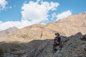 Hiker with backpack sitting on stone against rocks in bright sunlight. Alone man with photo camera in bright sun near sunlit rocky mountain wall. Tourist on moraines in high mountains in sunny day.