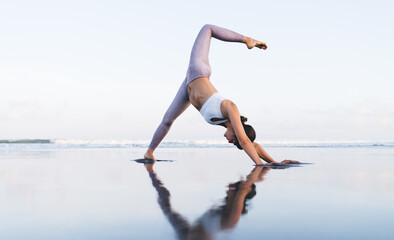 Flexible Caucasian female in active wear standing in asana with raised leg concentrated on keeping body shape, woman 20s enjoying yoga training at coastline feeling vitality inspiration and balance