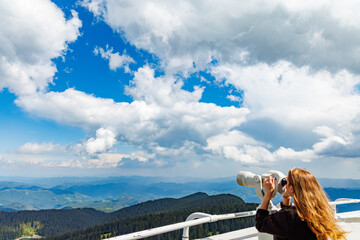 Girl tourist looks at landscapes of Rhodope Mountains and cloudy sky through telescope, on high...