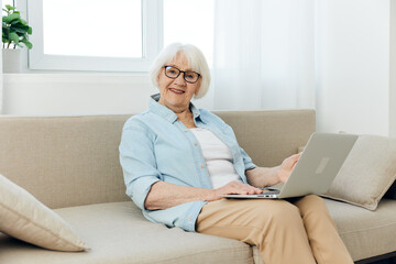 a sweet, joyful pensioner in a light shirt is sitting in a beautiful interior on a beige sofa near the window and holding a laptop on her knees, smiling broadly using the Internet