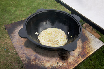 Onions are fried in a cast-iron cauldron. There is smoke and steam. Prepares food outdoors.