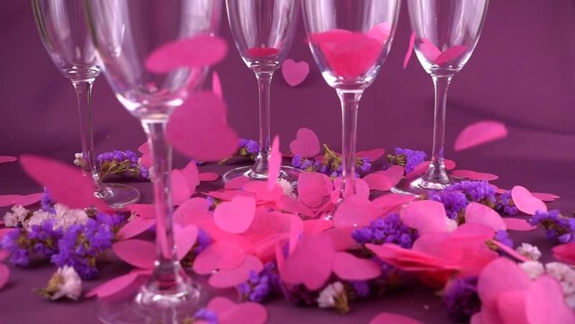 Falling heart shaped confetti on glasses wine and flowers on a purple background. Slow motion.