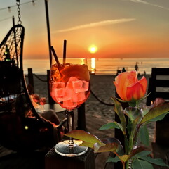  red roses and glass of orange  water  with ice  on wooden table top    sunset at sea  in beach restaurant view in pink sky and sea nature landscape