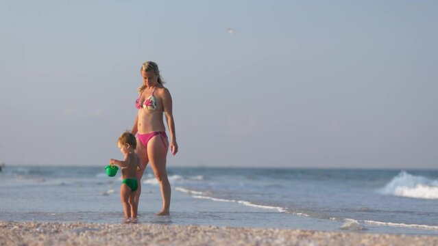 Mom walks along the sea beach with her son collecting seashells under the summer sun