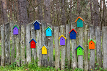 Wooden fence with decorative elements in the form of multicolored stylized birdhouses. Colored decorative birdhouses on a picket fence.