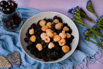 Frozen yellow raspberries and mulberries on a plate along with a jug of sweet cherries and flowers on a blue background